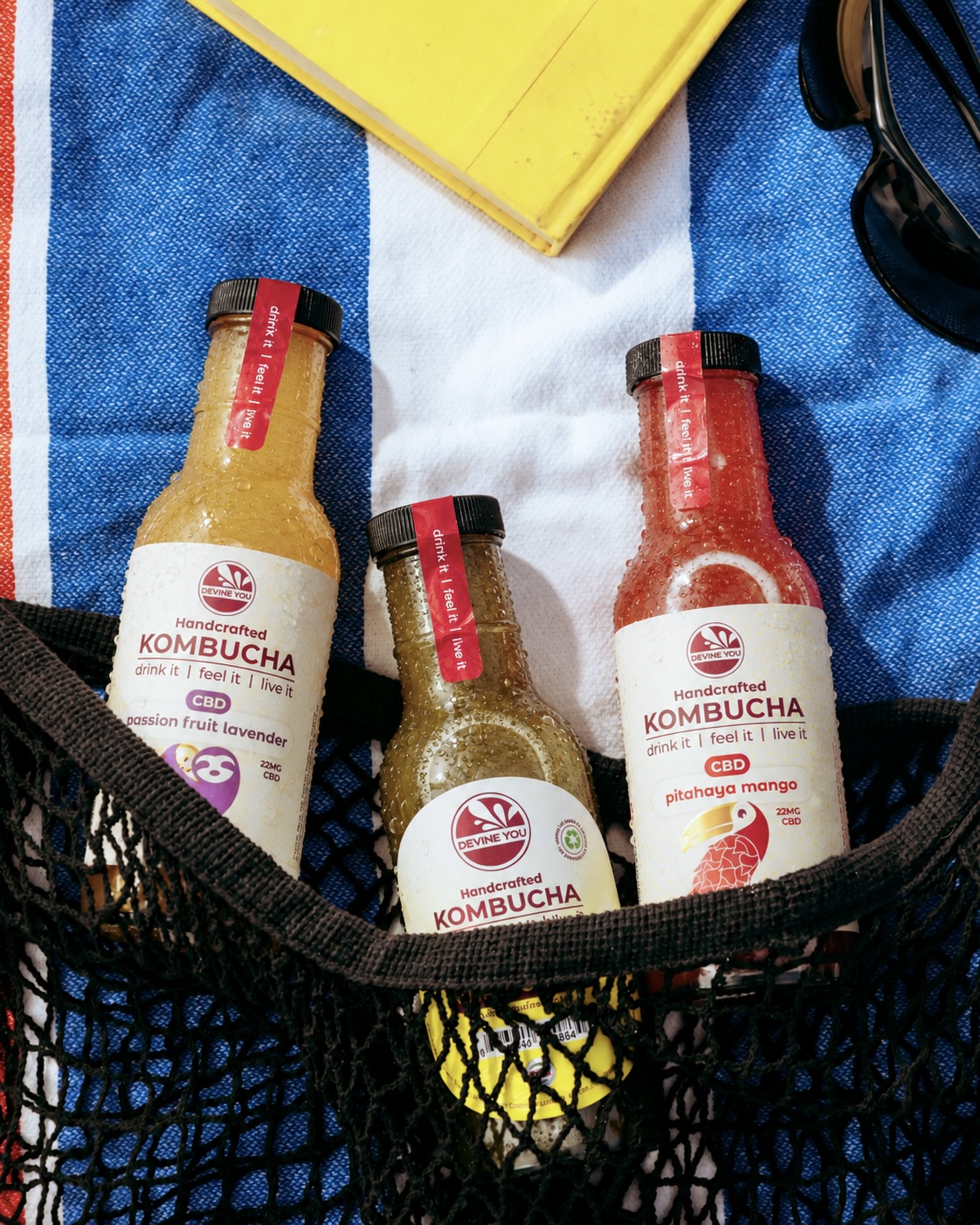 Three Devine You kombucha bottles in a mesh bag on a beach towel